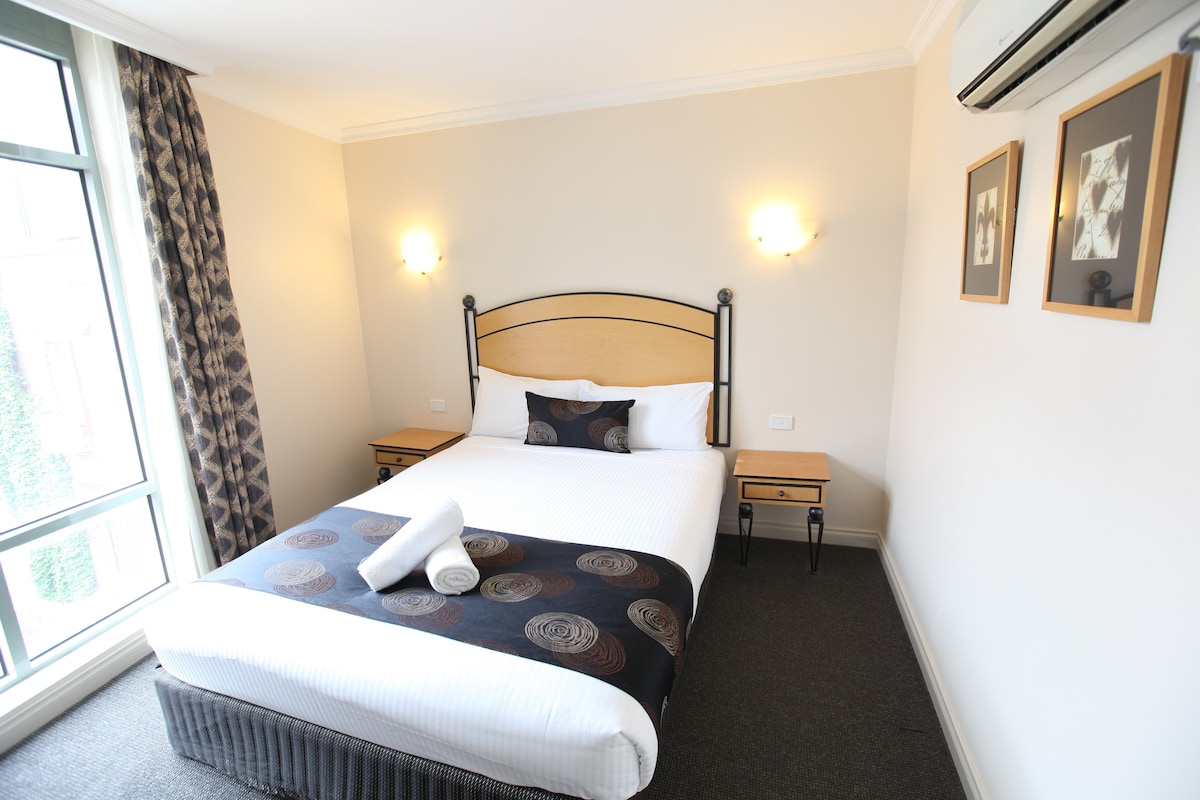 A well-appointed bedroom is highlighted by a spacious bed with a patterned blanket. Two bedside tables with lamps flank the bed, and the room features large windows allowing natural light. Framed artwork adorns the wall above, adding a touch of character.