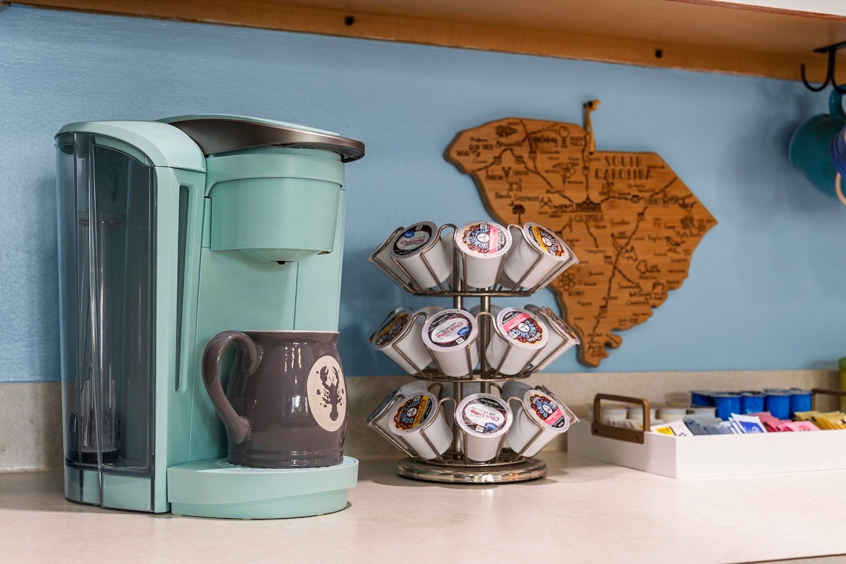 A teal coffee maker is positioned on a light countertop, with a grey mug placed beneath it. A rotating display holds an assortment of coffee pods. A wooden map of South Carolina is mounted on the wall behind, adding a local touch.