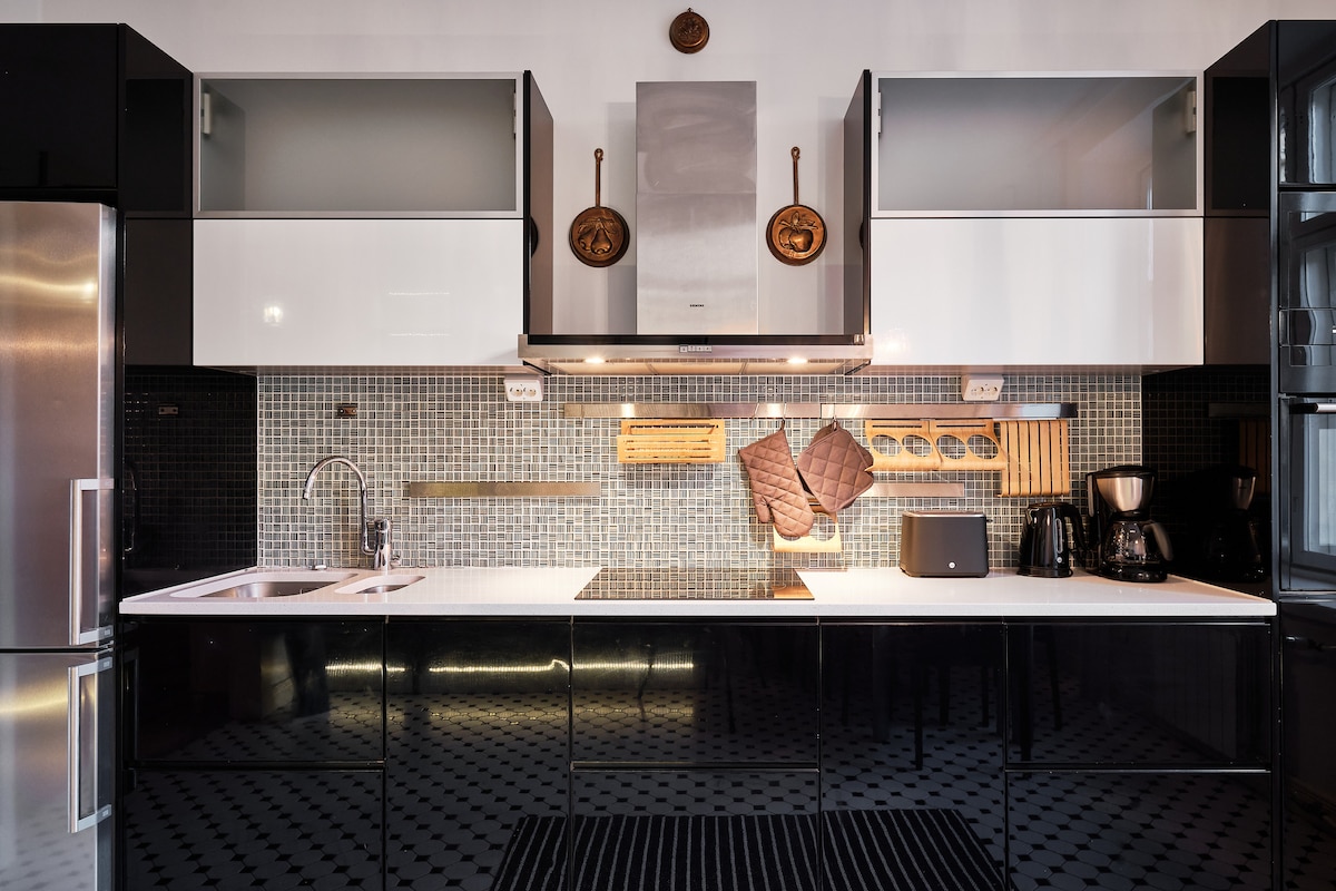 A modern kitchen is equipped with sleek black and white cabinetry, complemented by a stylish backsplash featuring patterned tiles. Stainless steel appliances and a coordinated countertop are visible, along with kitchen utensils neatly organized on a wooden rack, creating a practical cooking environment.