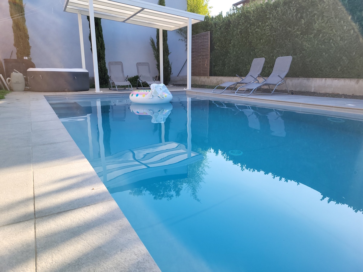 The outdoor pool is surrounded by a sleek stone deck, reflecting the clear sky. Two reclining lounge chairs are positioned nearby under a shaded pergola, creating a relaxed atmosphere. A float is situated in the water, enhancing the inviting nature of the space.