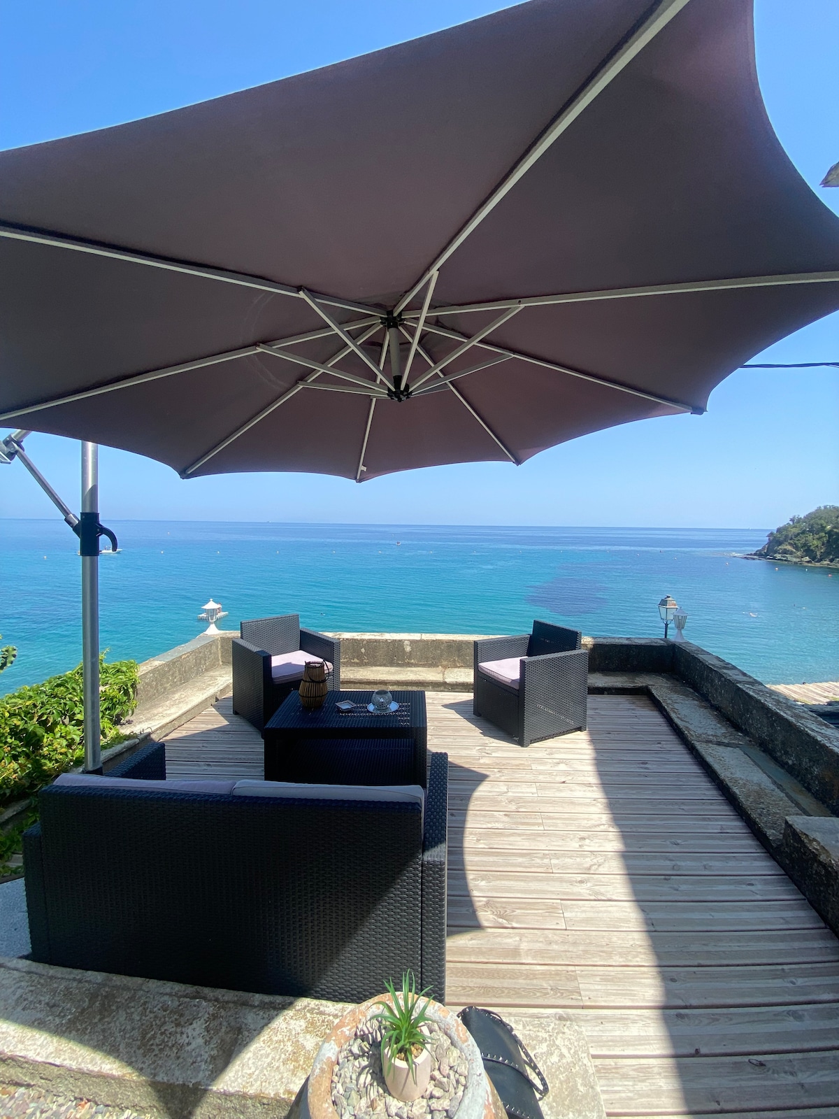 An exterior terrace features a shaded seating area with dark wicker furniture, facing the shimmering blue Mediterranean Sea. A large umbrella provides shade, and a small potted plant adds a touch of greenery to the inviting outdoor space.