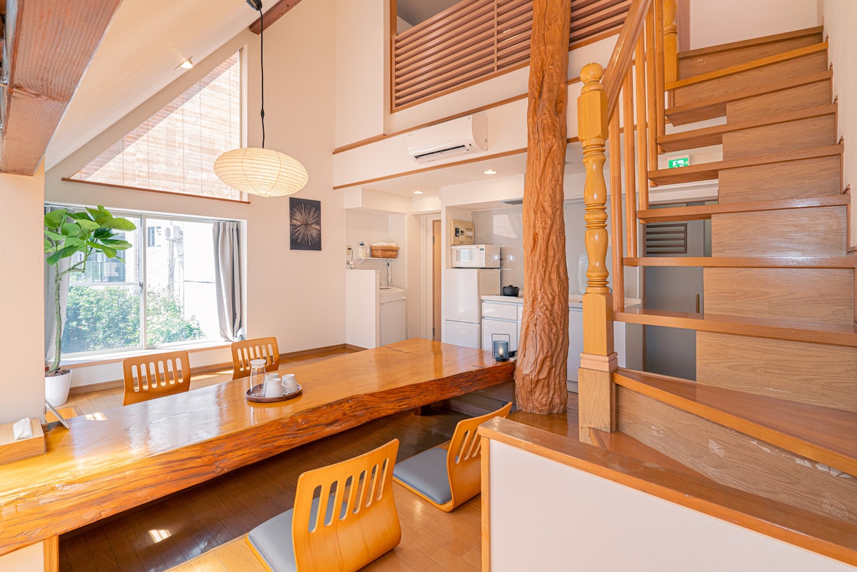An open-plan living space features a natural wood dining table surrounded by four wooden chairs. A cozy atmosphere is created by soft lighting and a partially visible kitchen in the background. A staircase leads to the upper loft area, enhancing the spaciousness.