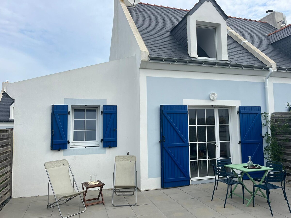 The exterior of the house features blue shutters and a welcoming entrance. Two loungers are positioned on the patio alongside a green table and chairs. The surroundings include a modest wooden fence and a small garden area, enhancing the outdoor space's appeal.