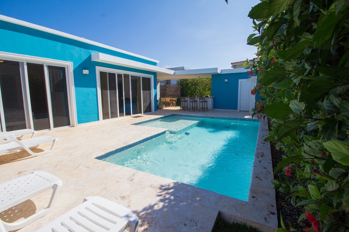 A bright blue exterior surrounds a spacious patio featuring a sparkling swimming pool. Lounge chairs are positioned nearby, alongside a shaded seating area with a table. Lush greenery borders the space, enhancing the outdoor ambiance.