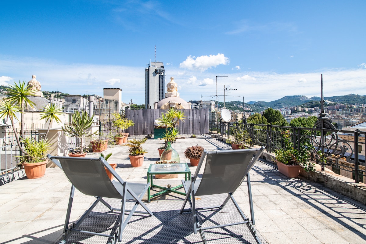 A panoramic terrace offers a relaxed seating area with two lounge chairs positioned towards a view of the cityscape. Potted plants and greenery surround the space, enhancing the outdoor ambiance against a clear blue sky.