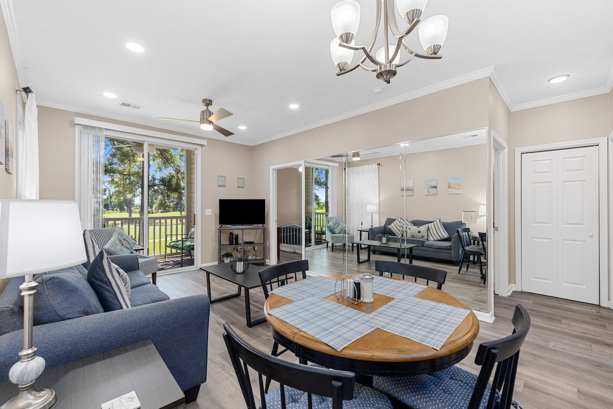 An open living and dining area features a circular wooden dining table with dark chairs and a blue table setting. The seating area includes a blue couch and armchair, with a TV visible in the background. Large windows offer views of the outdoors.