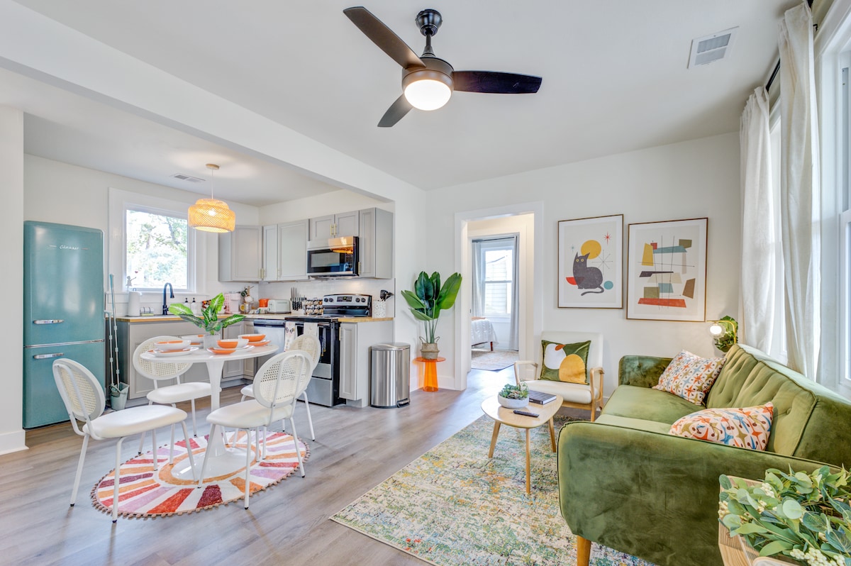 A bright and open living space is displayed, featuring a cozy green sofa and a round dining table set for four. A modern kitchen with stainless steel appliances is visible in the background, alongside large windows that allow natural light to fill the room.