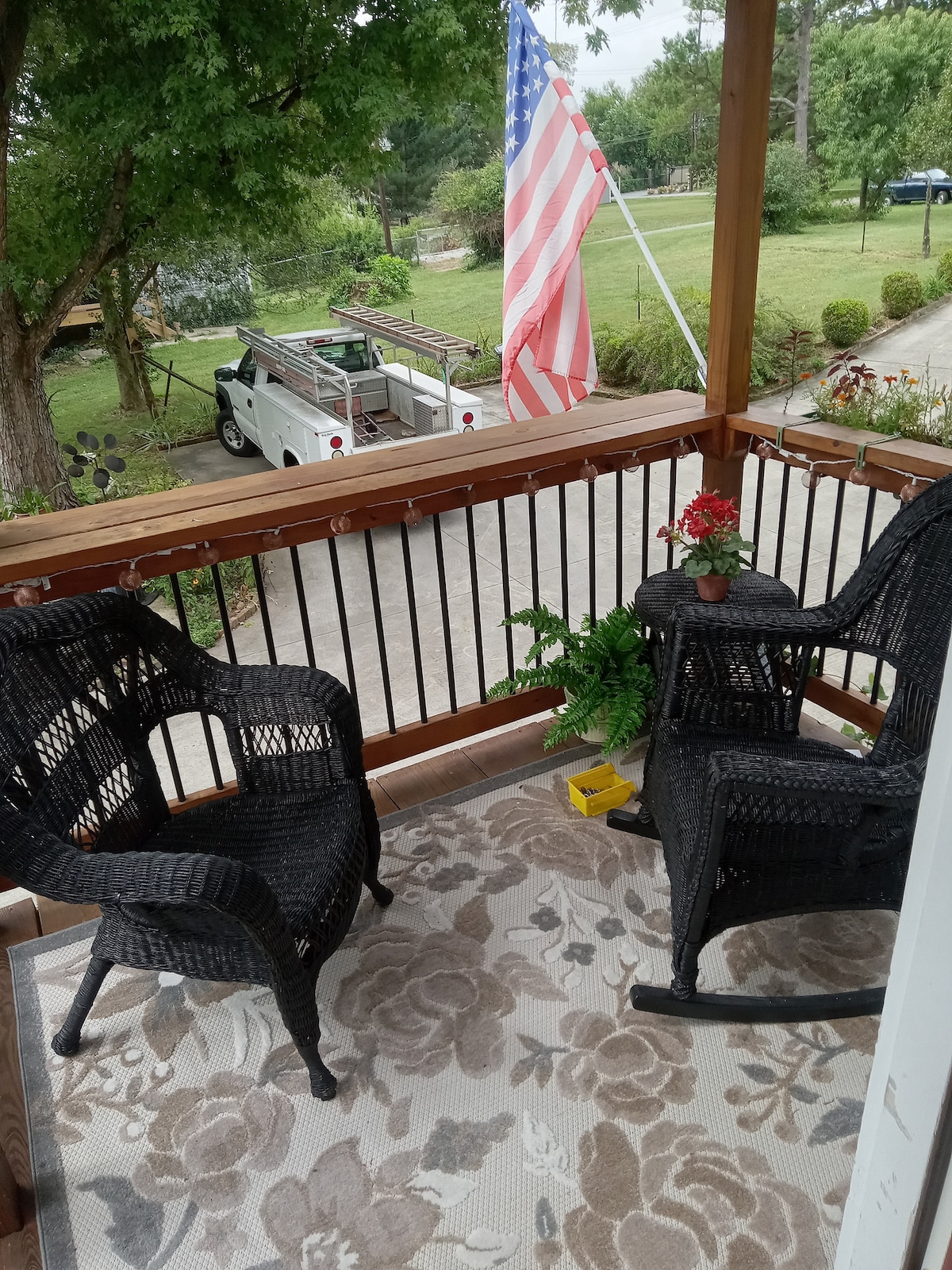 A covered balcony area features two black wicker chairs positioned around a small table. A potted flower adds a pop of color, while a patterned rug adds texture underfoot. An American flag is visible in the background, with greenery and a driveway just beyond.