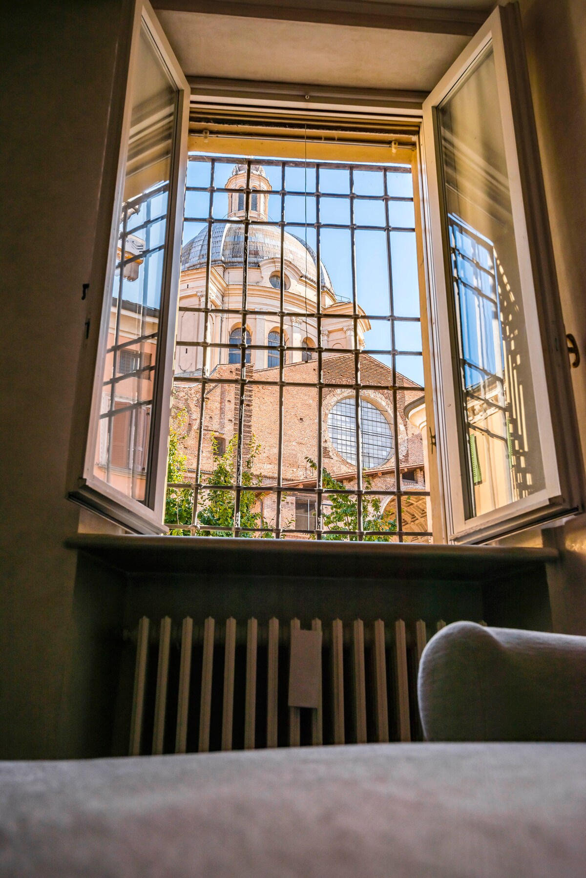 A scenic view is presented from an open window, framing the exterior of a historic church with a distinct dome. Light floods the space, highlighting the architectural details of the church and surrounding greenery. A radiator is visible below the window.