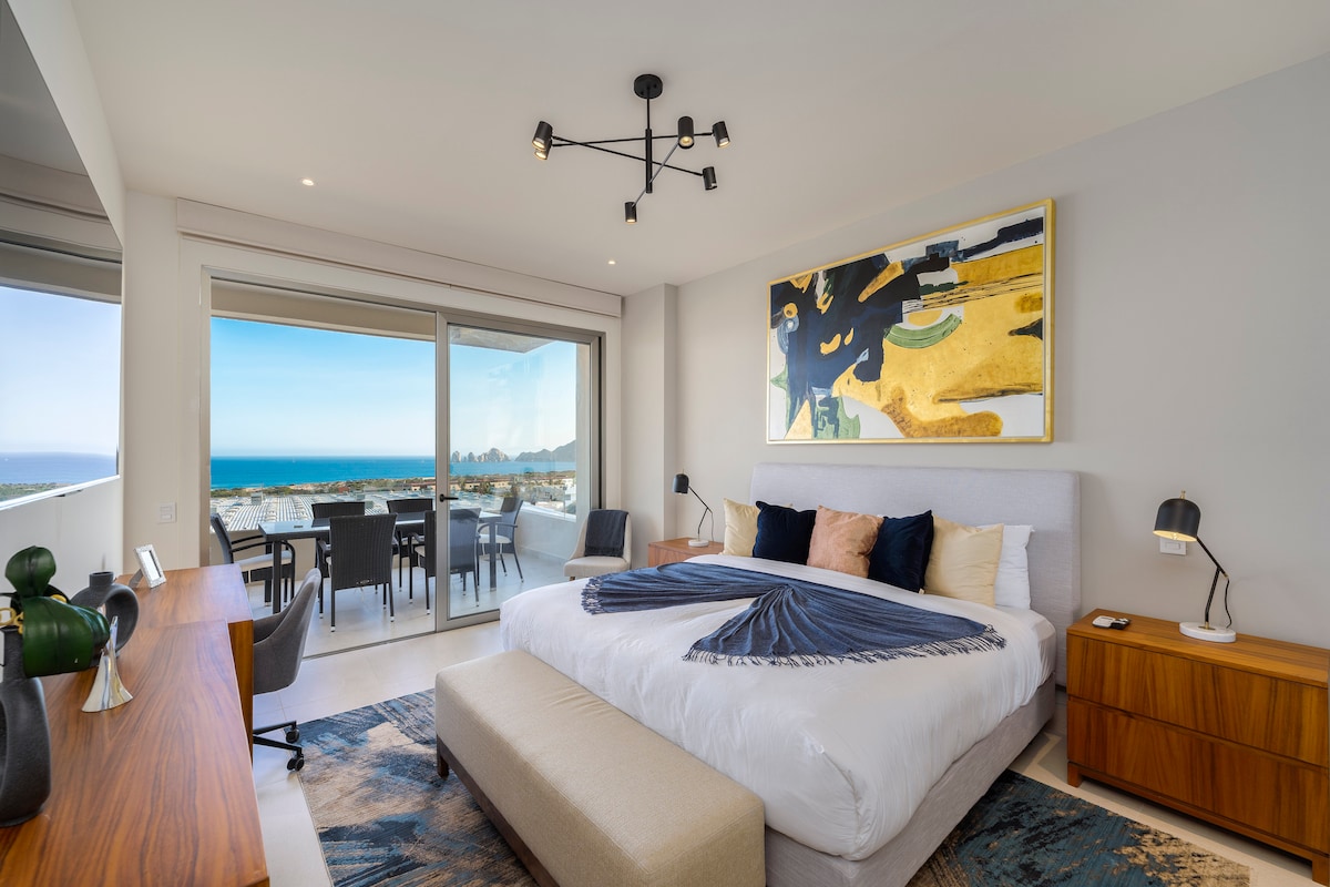 A modern bedroom features a spacious bed with layered bedding and decorative pillows. A distinct artwork is displayed above the bed. Floor-to-ceiling sliding doors provide a scenic view of the Bay, with a furnished balcony area visible. A functional desk and lamp are included.