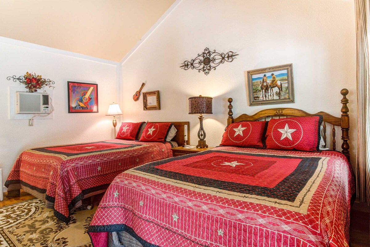 A sleeping area features two Queen Beds, each adorned with red and black patterned quilts. An artwork depicting horses hangs above the beds. A lamp and a small air conditioning unit are positioned on the side tables, with light-colored walls and wooden accents completing the space.