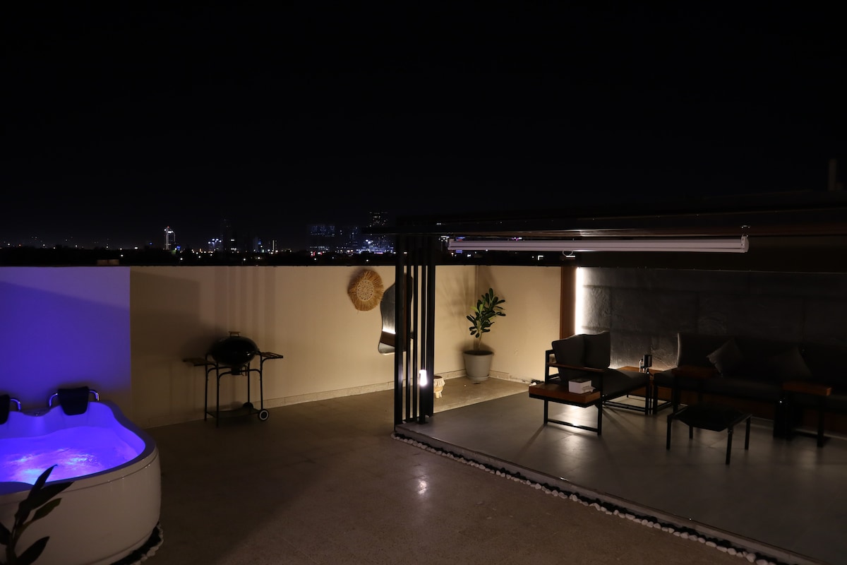An outdoor patio area features a jacuzzi with soft blue lighting, surrounded by modern seating arrangements. A grill is positioned nearby, with a potted plant adding a touch of greenery. The city skyline is visible in the distance, enhancing the tranquil evening atmosphere.