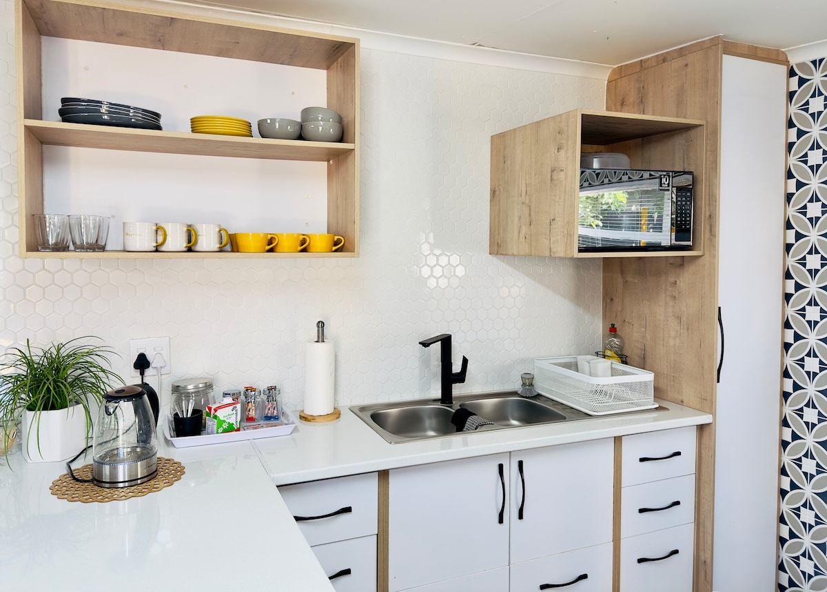 A modern kitchen is shown, featuring a blend of light wood and white cabinetry. Open shelves display an assortment of plates and mugs, while a sleek black faucet is positioned above a stainless steel sink. An organized countertop includes a kettle, dish rack, and microwave.