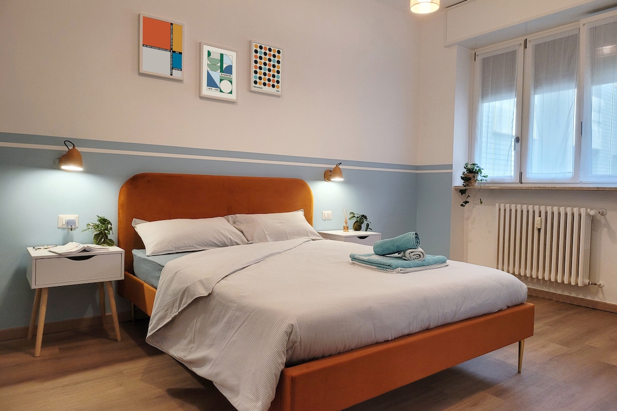 A modern bedroom features a spacious bed with a light-colored duvet and two pillows. Bedside tables with lamps are positioned on either side. Colorful artwork adorns the wall above the bed, and natural light enters through two windows, complemented by light curtains.