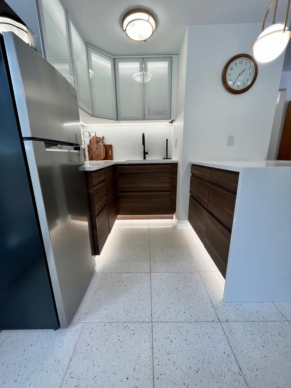 A modern kitchenette is showcased, featuring sleek dark cabinetry paired with a light countertop. Stainless steel appliances, including a refrigerator, enhance the space. Soft under-cabinet lighting highlights the clean lines and contemporary design, providing an inviting area for meal preparation.