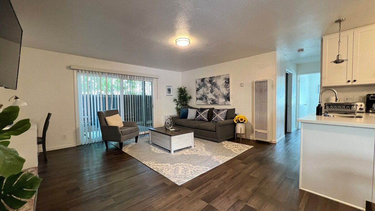 A spacious living area is depicted, featuring a comfortable gray sofa and an accent chair positioned beside a coffee table on a patterned area rug. Natural light filters through sliding glass doors, with indoor plants adding a touch of greenery. An open kitchen area is visible in the background.