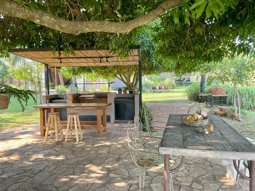An outdoor kitchen area features a wooden countertop and stools beneath a lush canopy of trees. A rustic dining table with chairs is situated nearby, surrounded by greenery, providing a serene spot for meals or gatherings.