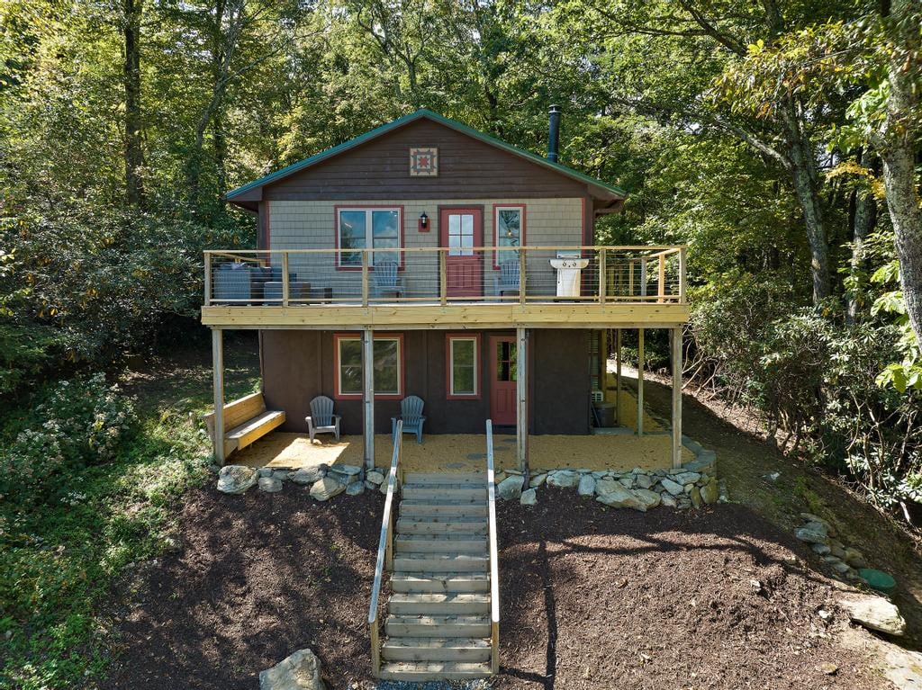 The cabin's exterior showcases a two-story structure nestled among trees, featuring a spacious deck with seating. Steps lead from the ground level to the deck, which is complemented by a rustic railing and a small table. The surrounding landscape includes a mix of greenery and boulders.