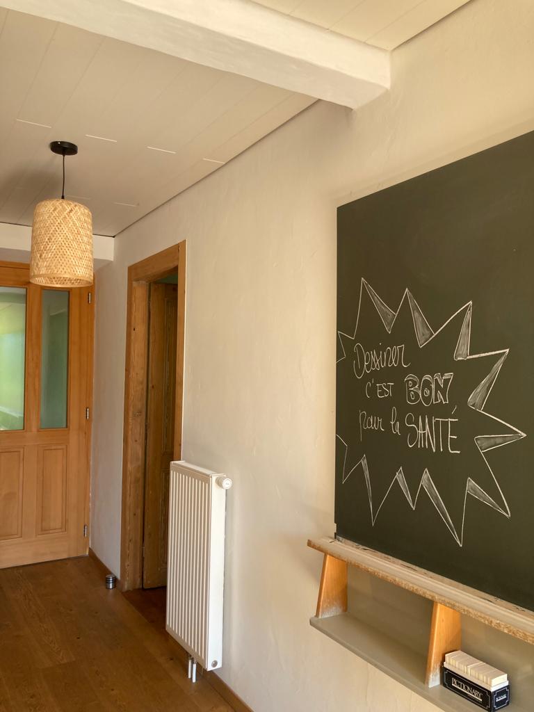 A hallway features a textured light fixture hanging from the ceiling. A chalkboard with a hand-written phrase is mounted on the wall, alongside a wooden shelf. A radiator is positioned below the chalkboard, and a glass-paneled door leads to another space.