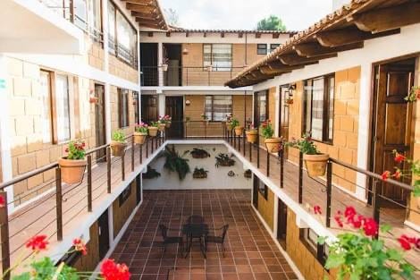 A central courtyard is surrounded by two-story accommodations, featuring warm-toned brick walls and wooden beams. Potted plants line the balcony railings, while a table and chairs are positioned on the tiled floor, inviting relaxation in a serene outdoor space.