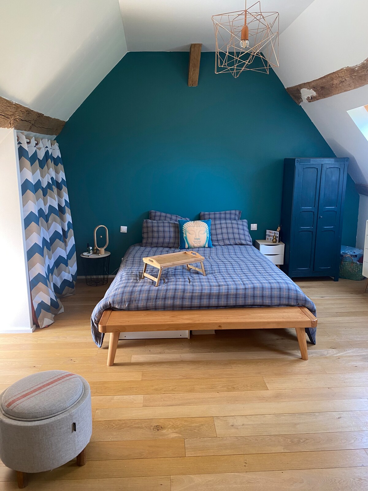 The spacious bedroom is characterized by a bold teal wall, complemented by wooden beams and warm wood flooring. A cozy bed with checkered bedding occupies the center, alongside a bedside table and a blue wardrobe. A patterned curtain adds texture to the bright space.