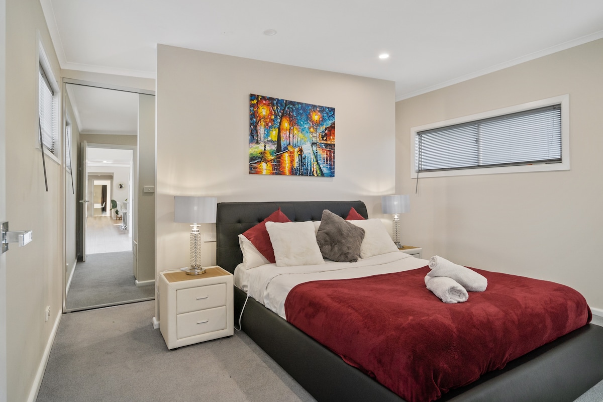 A cozy bedroom is presented with a spacious bed featuring bold red and white bedding. Soft gray carpeting covers the floor, while a vibrant piece of artwork adorns the wall above the headboard. Two bedside lamps provide illumination, and a mirror reflects the room's design.