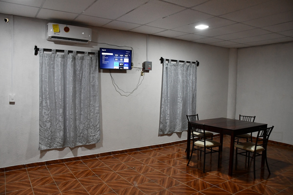 A dining area features a rectangular wooden table surrounded by four black chairs. Two large windows with gray curtains allow natural light, and a wall-mounted television is positioned across from the table. An air conditioning unit is visible in the ceiling.