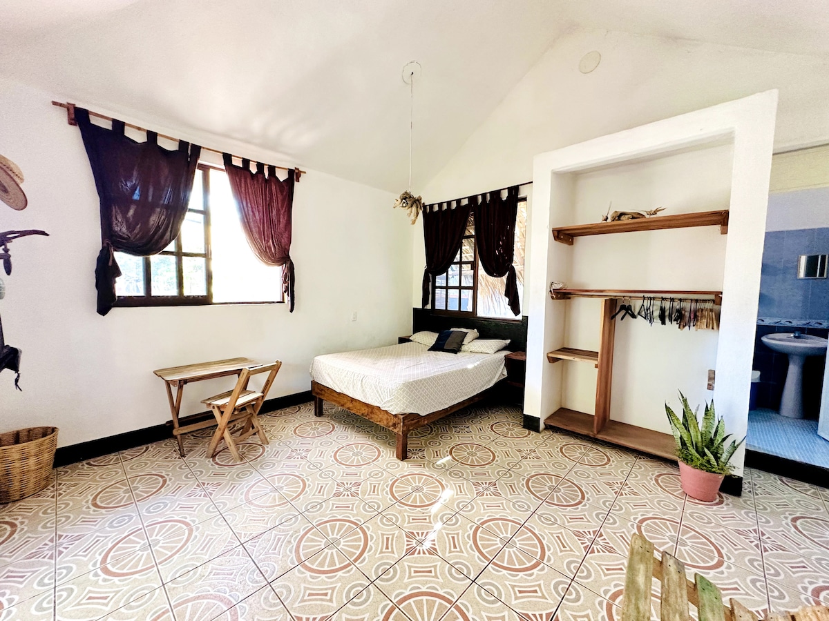 The room features a spacious bed positioned near large windows adorned with dark curtains. A small wooden table and foldable chairs are placed in one corner. Decorative shelving and a plant are visible, with patterned floor tiles adding character to the space.