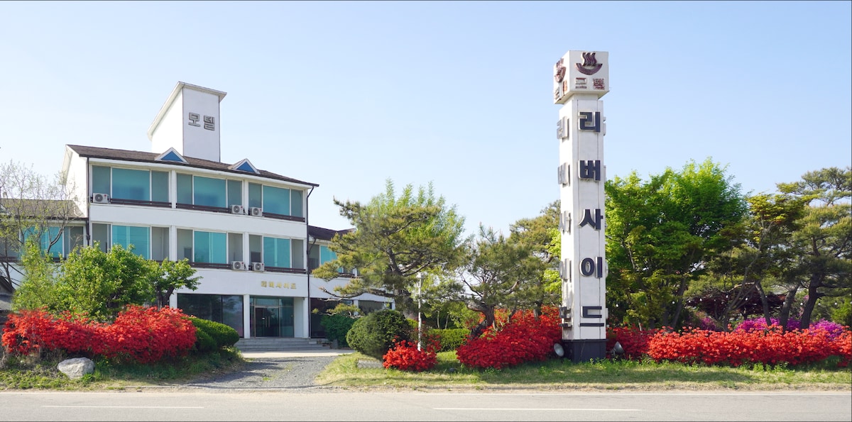 The exterior of a multi-story building is shown, characterized by large windows and a modern architectural style. A prominent sign displays the name of the establishment. Vibrant red and green landscaping surrounds the property, providing a refreshing contrast against the clear blue sky.