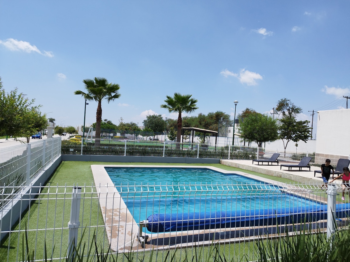 A sparkling pool is enclosed by a white fence, surrounded by lush green grass and palm trees. Several lounge chairs are visible along the poolside, providing a relaxing space. Clear blue skies and bright sunlight enhance the refreshing ambiance of the area.