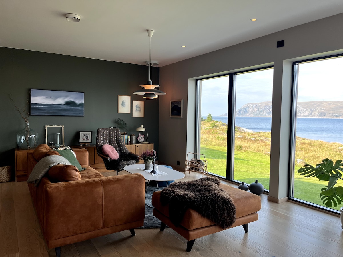 A modern living area features a brown leather sectional sofa and a matching chaise lounge. Large windows provide a scenic view of the coastline and greenery. The room is complemented by warm lighting and artistic decor, creating a stylish and comfortable space.