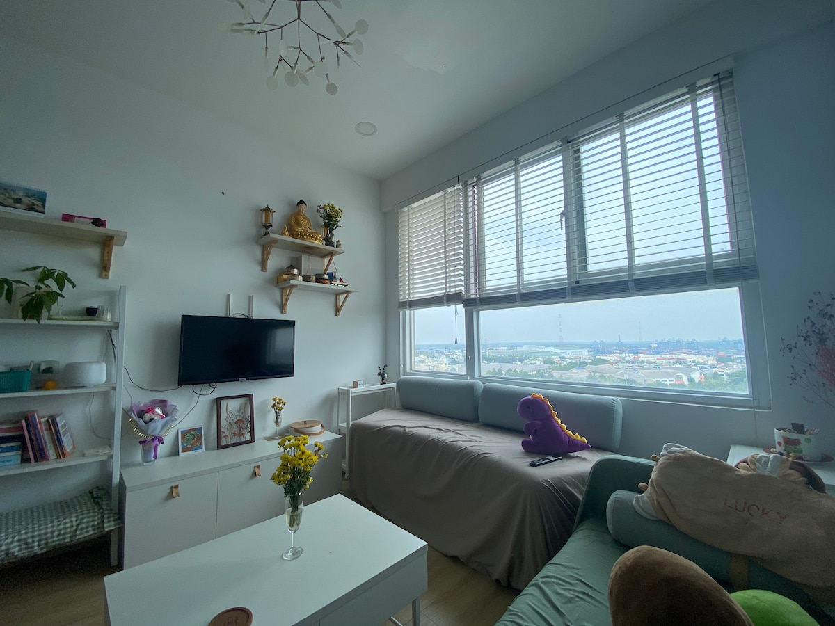 A bright living area features large windows with partially closed blinds, revealing a view of the surroundings. A TV is mounted on the wall, and floral arrangements adorn a white coffee table. Two cozy seating areas are complemented by decorative shelves and soft cushions.