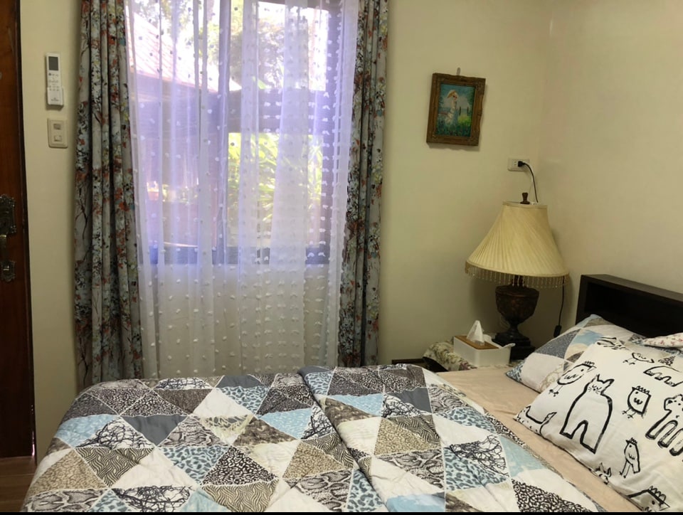 A cozy bedroom features a spacious bed with a patterned quilt and decorative pillows. Natural light filters through sheer curtains, illuminating the room. A bedside lamp and framed artwork add to the decor, while a window offers a view of the outdoors.