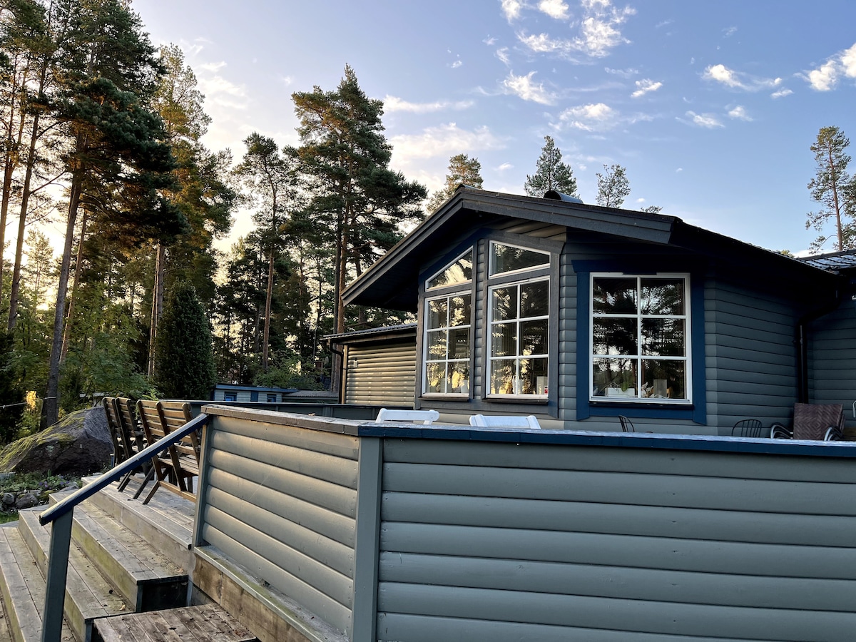 A tranquil wooden cabin features large windows that allow natural light to fill the interior. The deck is lined with comfortable seating, surrounded by trees that create a serene outdoor environment. The soft colors of the sky at sunset add to the calm setting.