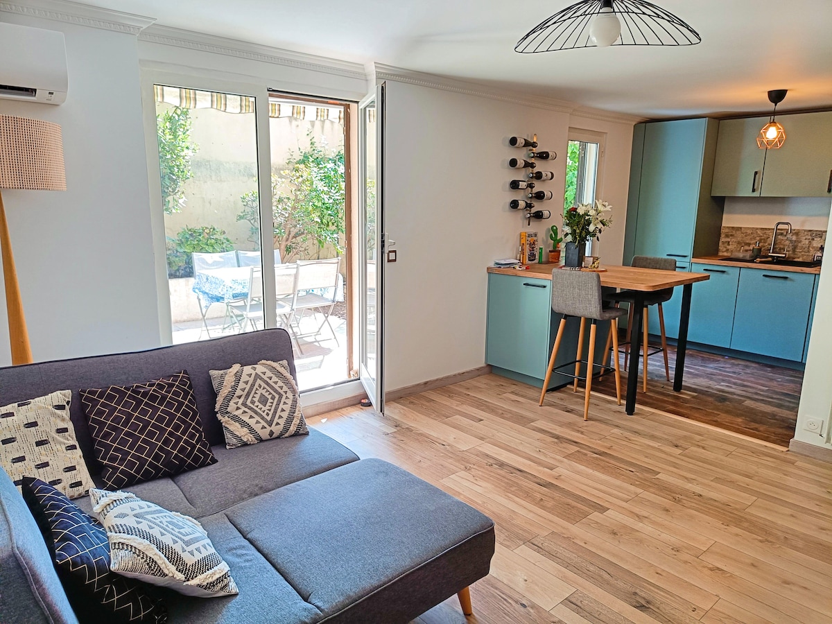 A modern open-concept living area features a comfortable sectional sofa adorned with patterned throw pillows. Natural light floods the space through large glass doors, leading to a private garden. A minimalist kitchen with blue cabinetry and a wooden dining table is visible in the background.