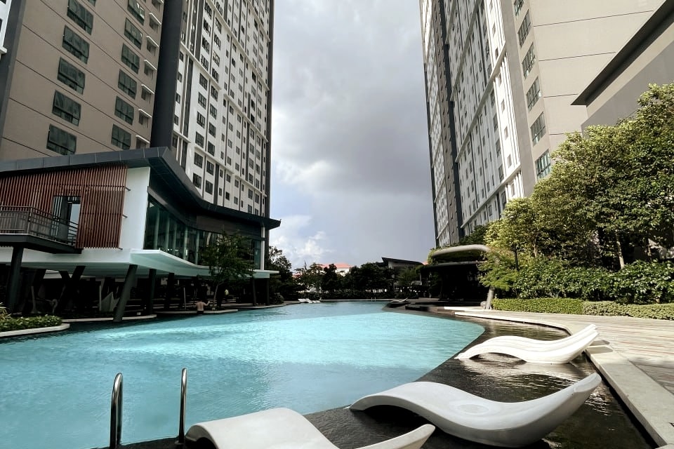 A large outdoor pool is surrounded by modern apartment buildings, featuring lounge chairs positioned beside the water. Lush greenery is present along the edges, and a clear sky with clouds is visible above, creating a spacious and refreshing atmosphere.