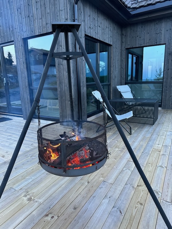 A hanging fire pit is situated on a wooden deck, surrounded by panoramic glass doors. Flames flicker within the mesh enclosure, providing warmth and light. Nearby, a comfortable chair and a second chair are positioned, offering an inviting space for relaxing outdoors.