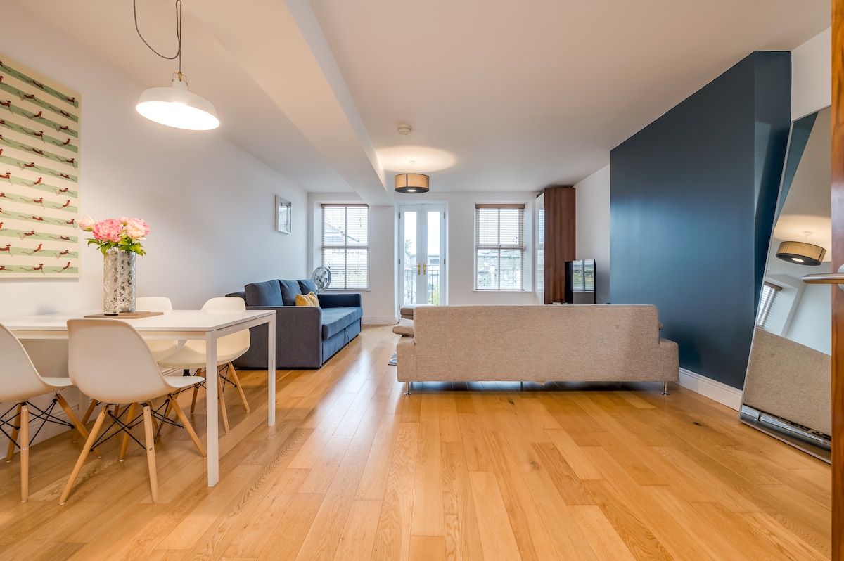 A large open-plan living area features wooden flooring and high ceilings, creating a spacious atmosphere. A grey sofa faces a flat-screen TV, while a dining table with white chairs is nearby. Natural light fills the space through large windows, enhancing the modern decor.