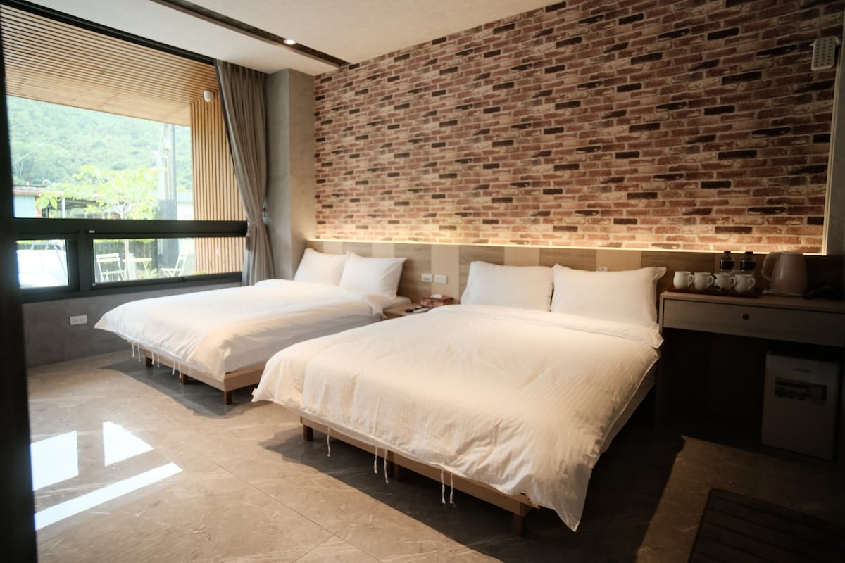 The sleeping area features two spacious beds with white bedding, complemented by a textured brick accent wall. Natural light floods in from a large window, which overlooks the garden. A small desk and mini-fridge are also present, enhancing the comfort of the space.