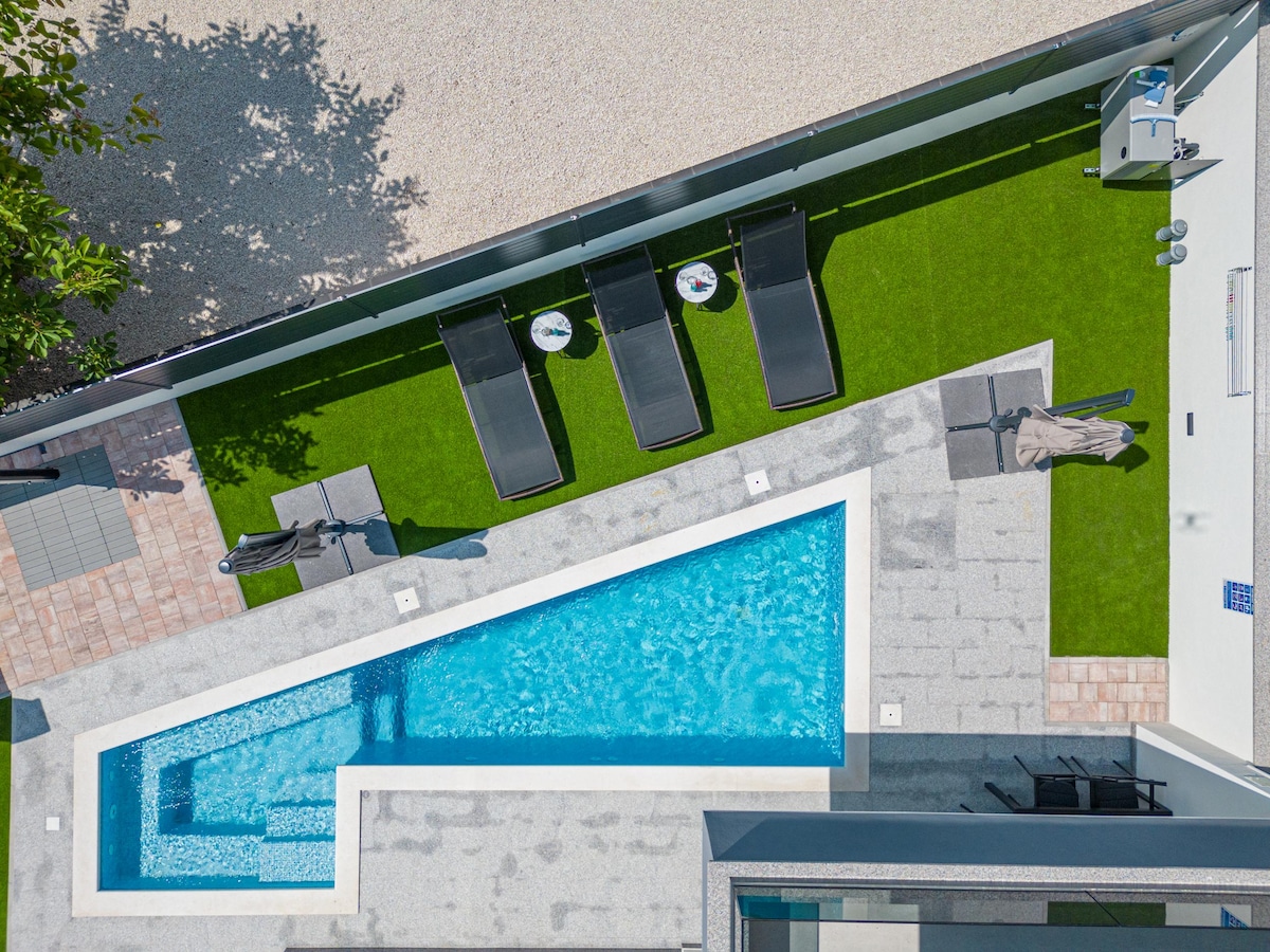 A private heated pool is surrounded by a landscaped area featuring lush grass and modern sun loungers. An outdoor dining space is visible, complemented by a charcoal grill nearby. The clean lines of the pool and patio create a serene outdoor retreat.