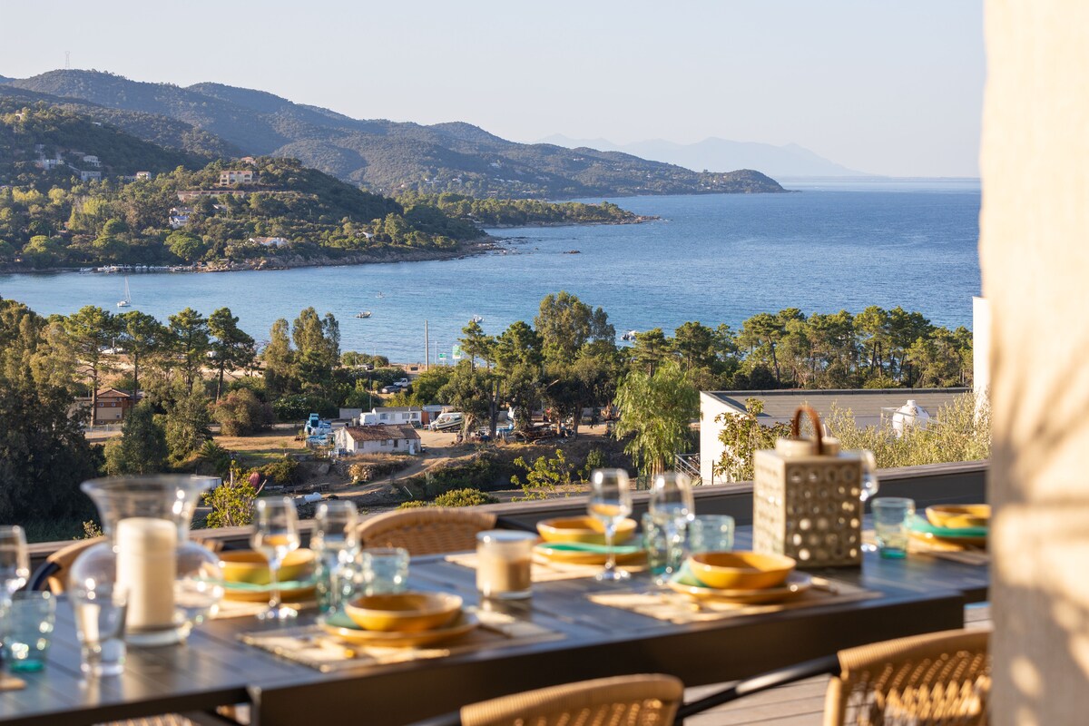 A dining table is set for a meal with colorful plates and clear glasses, overlooking a scenic coastal view. The distant hills and expansive ocean create a serene backdrop, enhancing the outdoor dining experience.