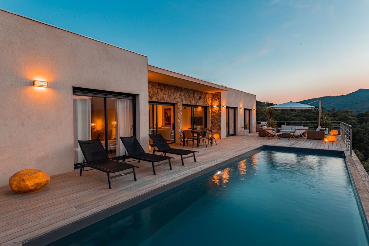 An outdoor area features a modern pool bordered by wooden decking. Several lounge chairs are positioned beside the pool, while an outdoor seating area with a large umbrella is visible in the background. The villa's large glass doors create a seamless connection between indoor and outdoor spaces.