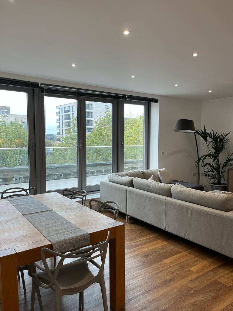 A bright living area features large glass doors leading to a balcony with views of greenery. A modern sofa faces a wooden dining table with four chairs, complemented by a floor lamp and indoor plants, contributing to a fresh, inviting ambiance.