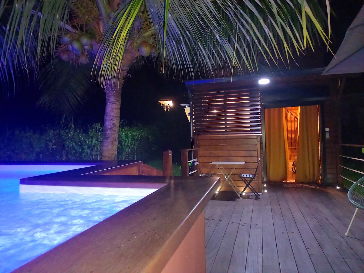 A private terrace is illuminated by ambient lighting, featuring a pool edged by a wooden deck. A palm tree provides shade, while an entrance to the bungalow is visible, partially framed by wooden slats.