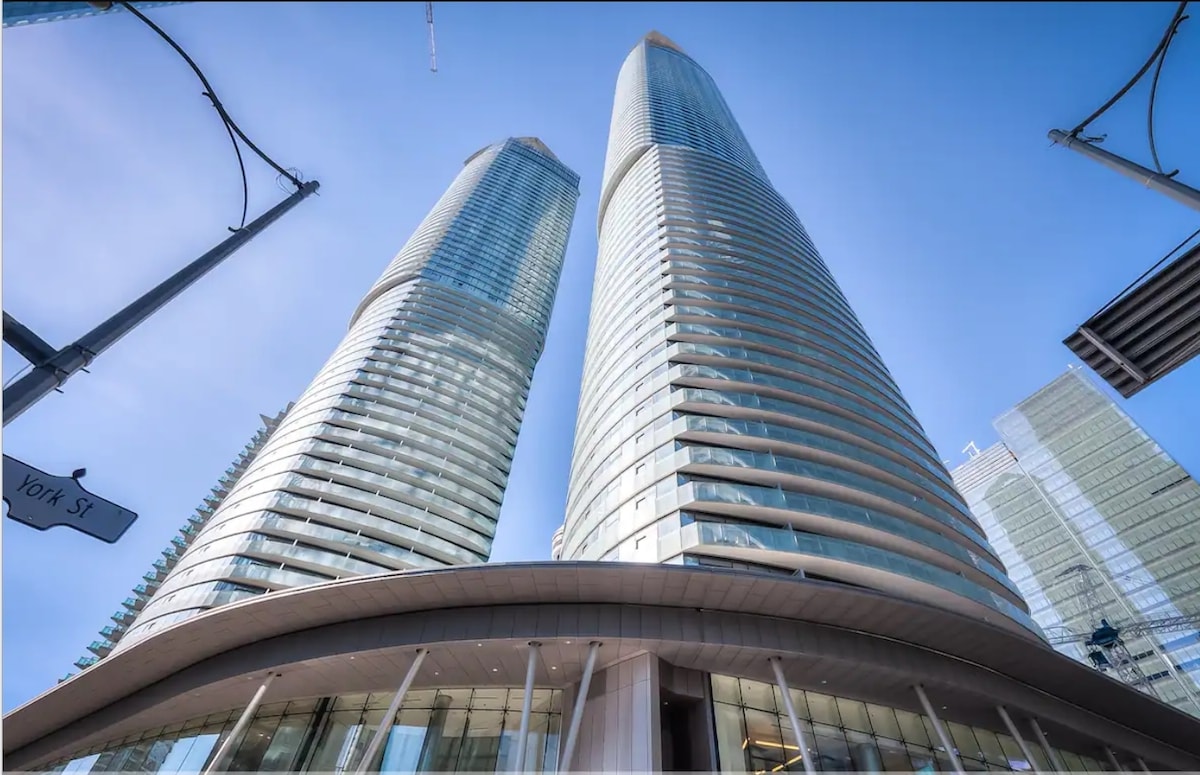 The modern condominium building is showcased from below, featuring sleek glass exteriors and curved architectural lines. The two towering structures rise into a clear blue sky, surrounded by urban elements that highlight downtown Toronto's vibrant atmosphere.