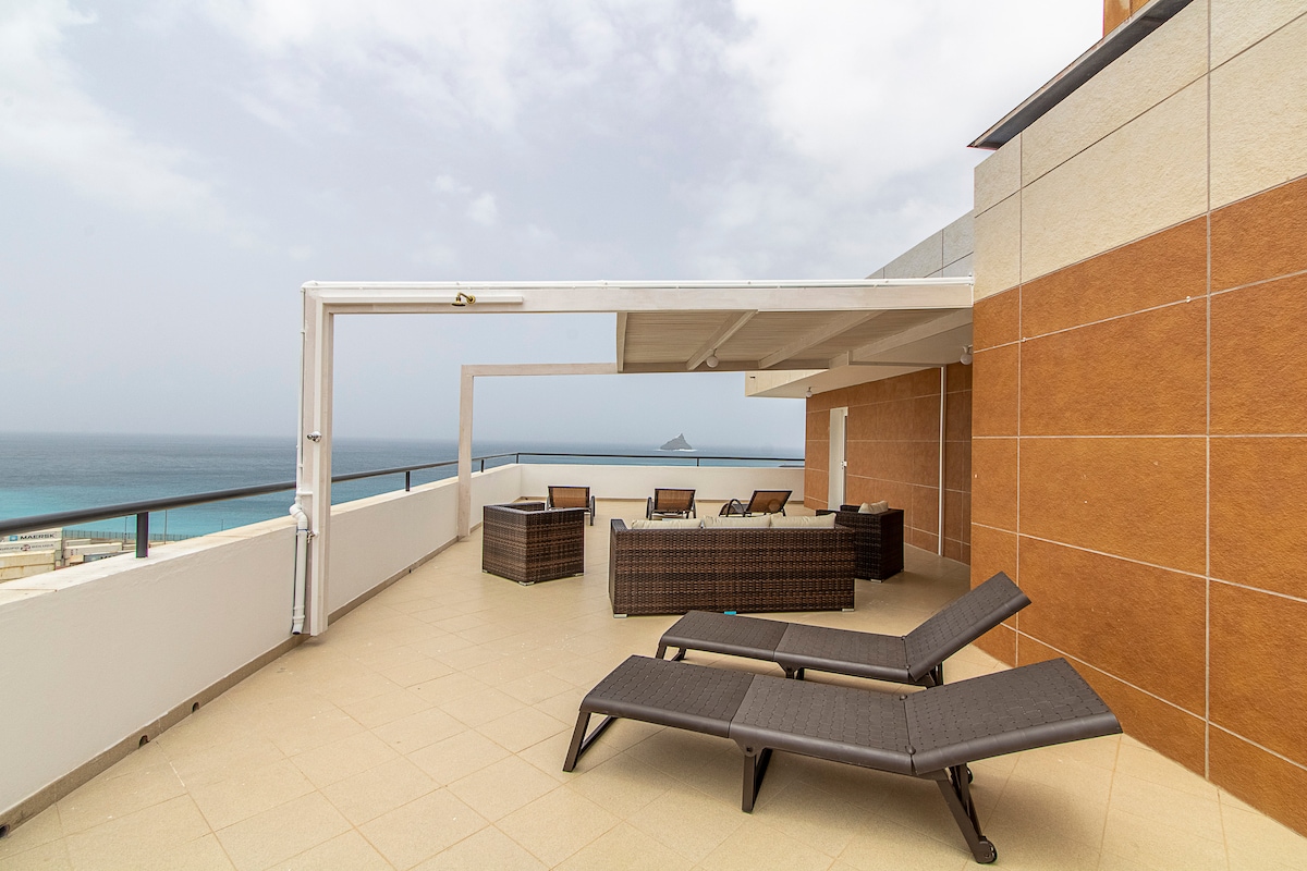 A spacious terrace is furnished with several lounge chairs and a shaded seating area. Neutral tiles cover the surface, while panoramic ocean views extend toward the horizon. A calming atmosphere is enhanced by the breezy open space.