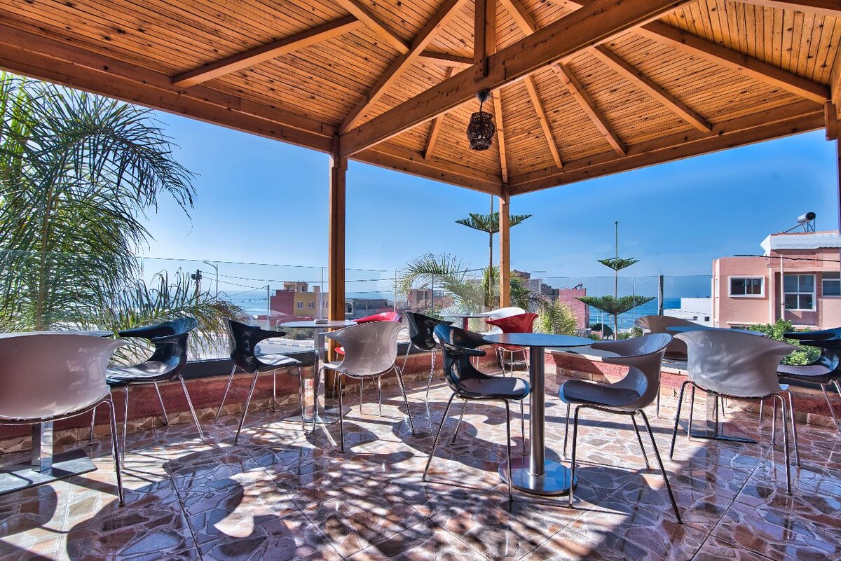 A covered terrace features a rustic wooden ceiling and is furnished with several modern chairs and tables. Potted plants add greenery, while views of the village and Atlantic Ocean are visible in the background, enhancing the inviting outdoor ambiance.