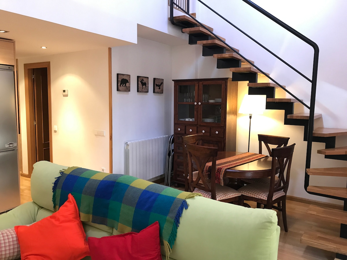 A spacious living area features a soft sofa adorned with colorful pillows. A wooden dining table sits nearby, surrounded by four chairs. An elegant staircase leads to an upper level, while art pieces are displayed on the wall, contributing to a cozy atmosphere.