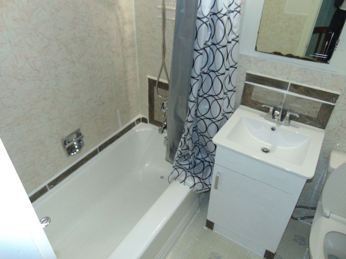 A compact bathroom features a white bathtub with a showerhead and a gray circular patterned curtain. A small white vanity with a sink complements the space, alongside light-colored walls and tile flooring. A mirror is positioned above the sink, enhancing natural lighting.