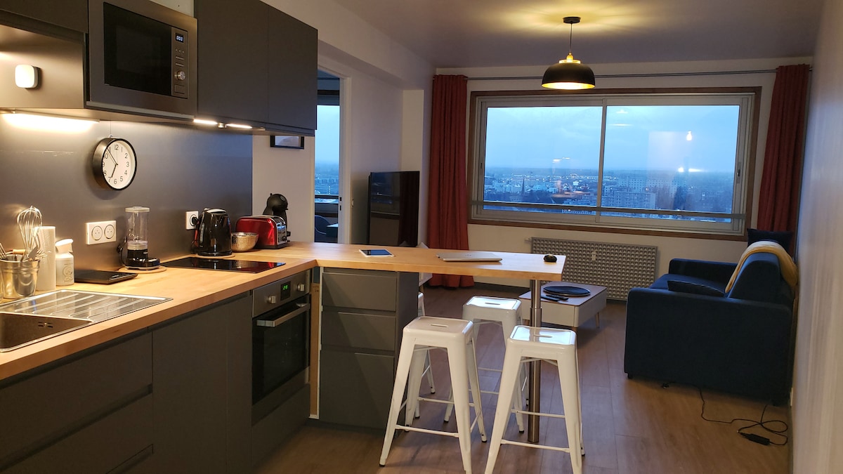 The kitchen area features modern appliances and an open layout, with a light wood countertop and dark cabinetry. Two metal stools are positioned at the counter, and a panoramic window reveals a city view, with a cozy seating area visible in the background.